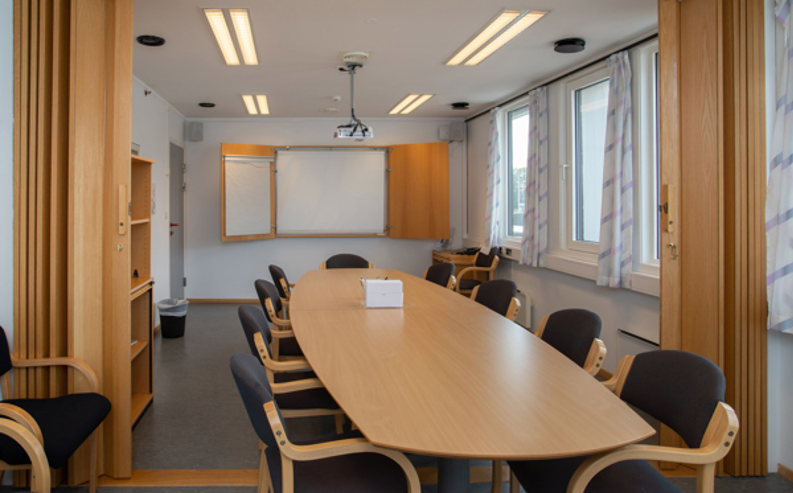 Møterom med ovalt bord i lyst tre og stoler i lyst tre med sort stoff rundt bordet. En tavle henger i enden av rommer.