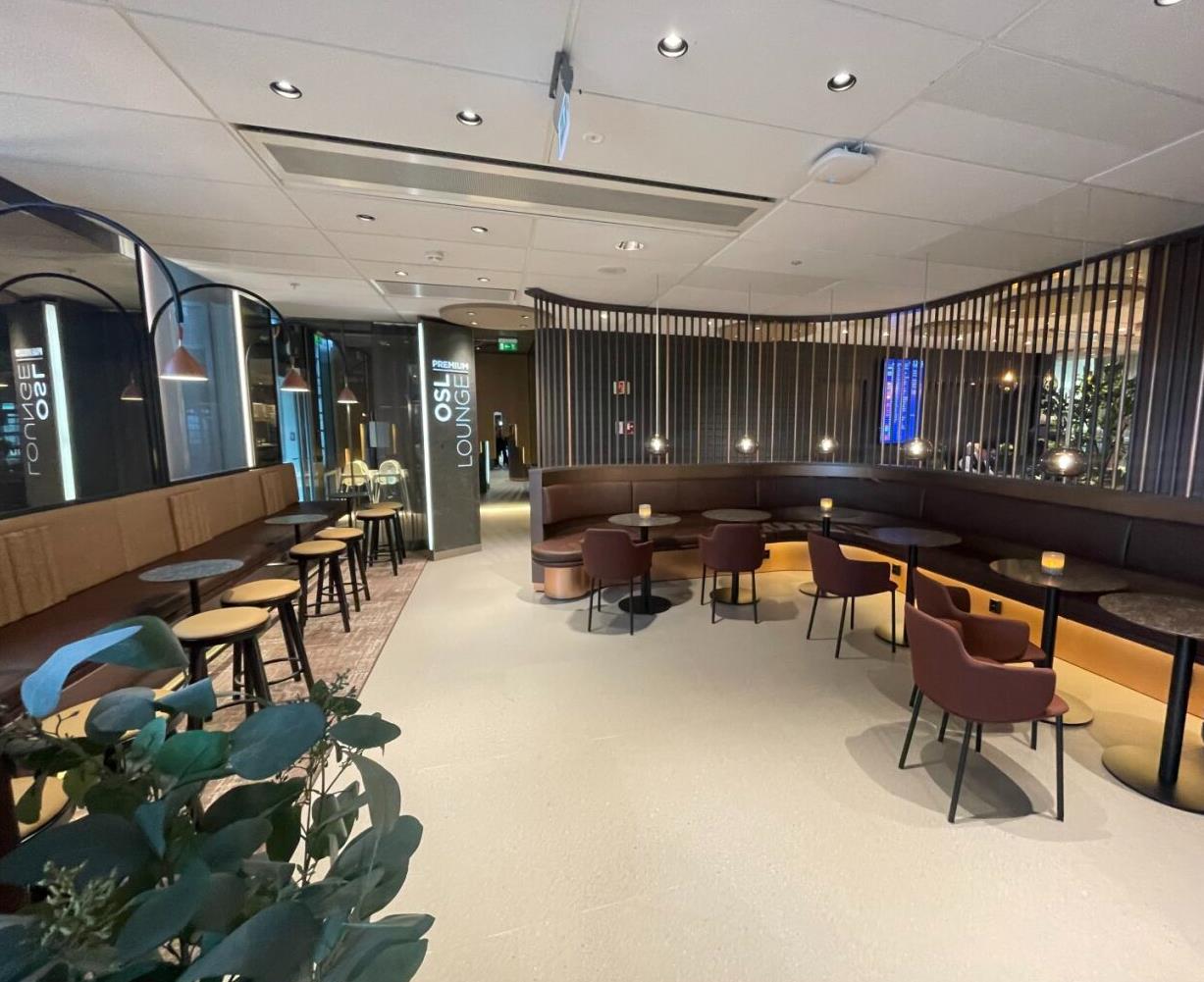 Spacious airport lounge interior with modern design, featuring brown seating, round tables, and ambient lighting, providing a comfortable waiting area for travelers