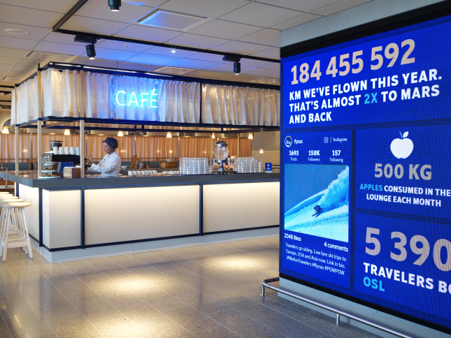 Cafe area in airport lounge with barista making coffee; digital display shows flight statistics, including number of travelers and amount of apples consumed.