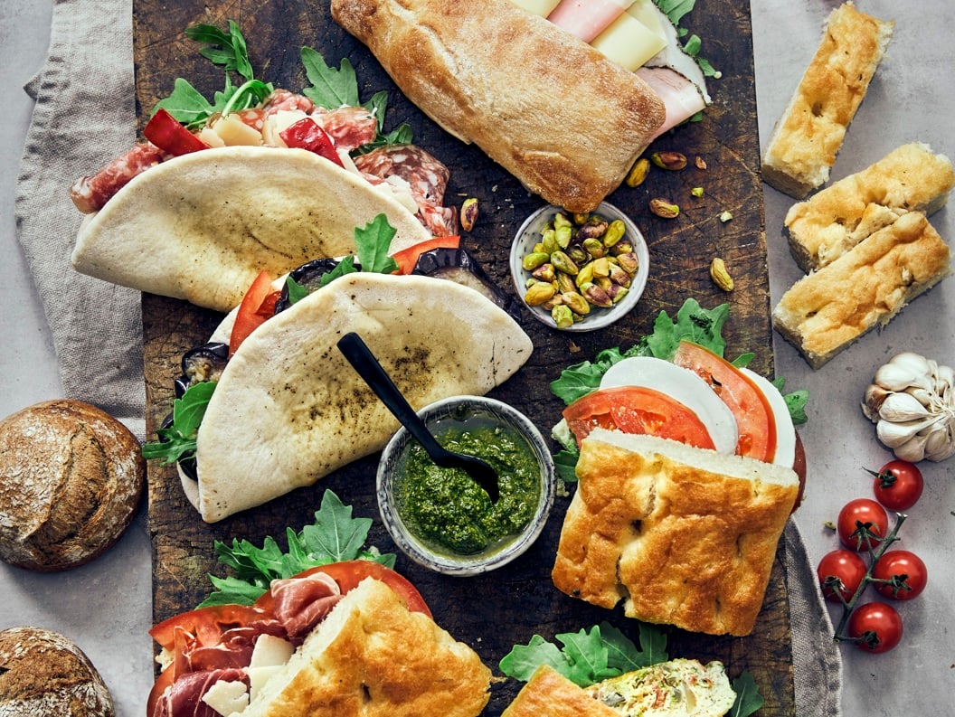 Italienske sandwicher med focaccia og ciabatta, fylt med spekemat, ost og grønnsaker. Serveres med pesto, aioli, nøtter og ferske cherrytomater. Matopplevelse på et trebord