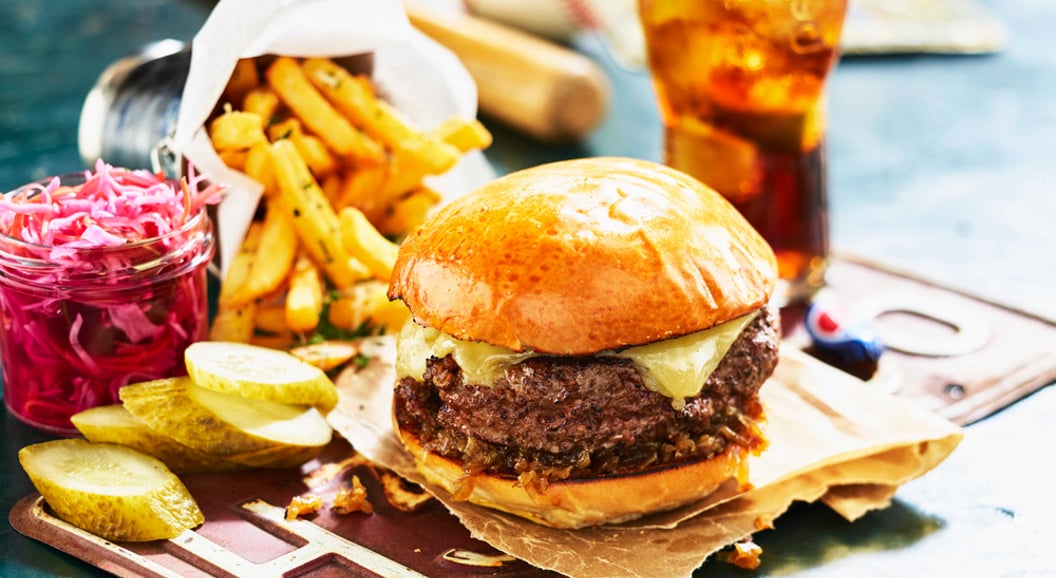 Saftig cheeseburger med sprø pommes frites, sylteagurk og rød coleslaw, ved siden av et forfriskende glass brus