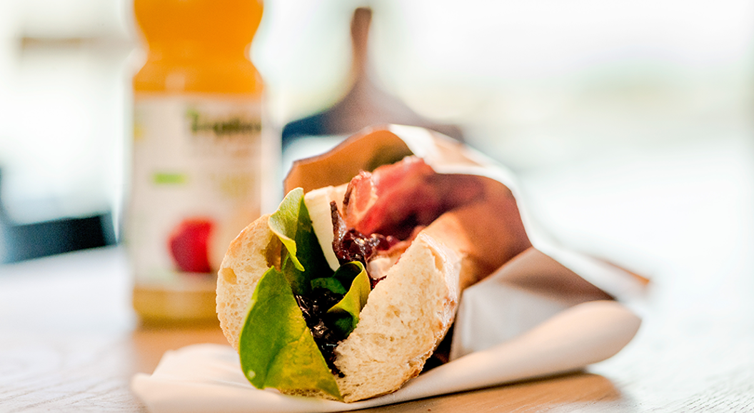 Freshly made sandwich with lettuce, bacon, and cheese on a wooden table, accompanied by a bottle of orange juice in the background