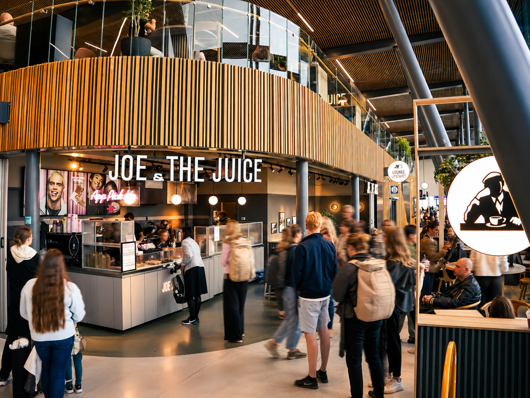 Assorterte Joe & The Juice-produkter, inkludert smørbrød, croissanter, grøt, juicer, açaí-boller og kaffe på en hvit bakgrunn med rosa emballasje og flasker med stille vann