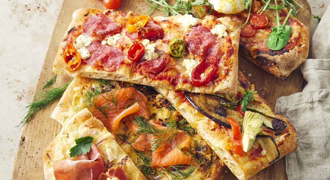 Assorterte gourmetflatbrødpizzaer på et trefat med toppings som inkluderer prosciutto, røkt laks, ruccola og ferske tomater, ledsaget av dippesauser på siden