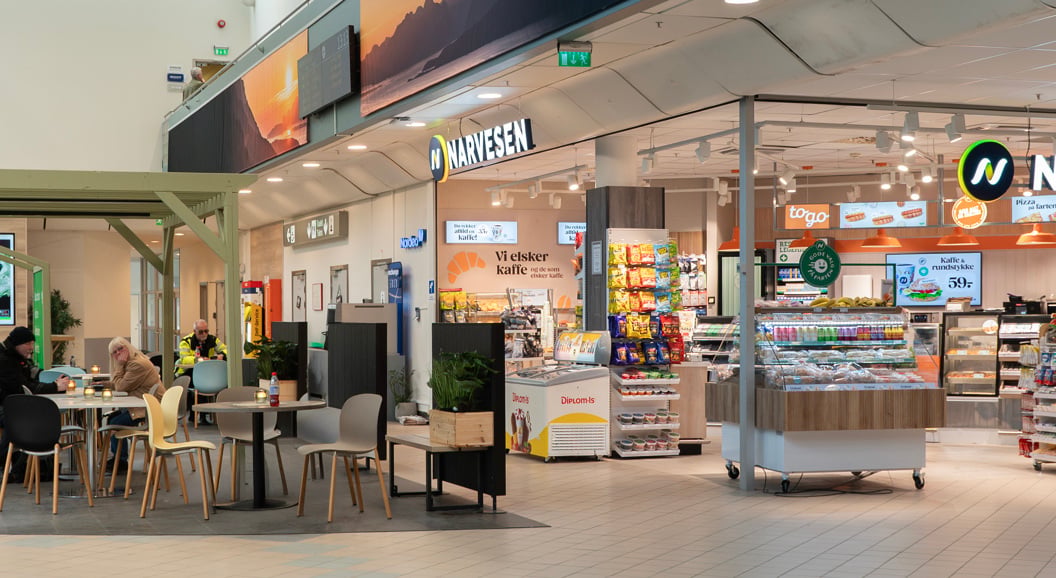 Moderne flyplasskiosk og kafé med sittende kunder, med Narvesen-branding, produktdisplayer og ankomstskilt