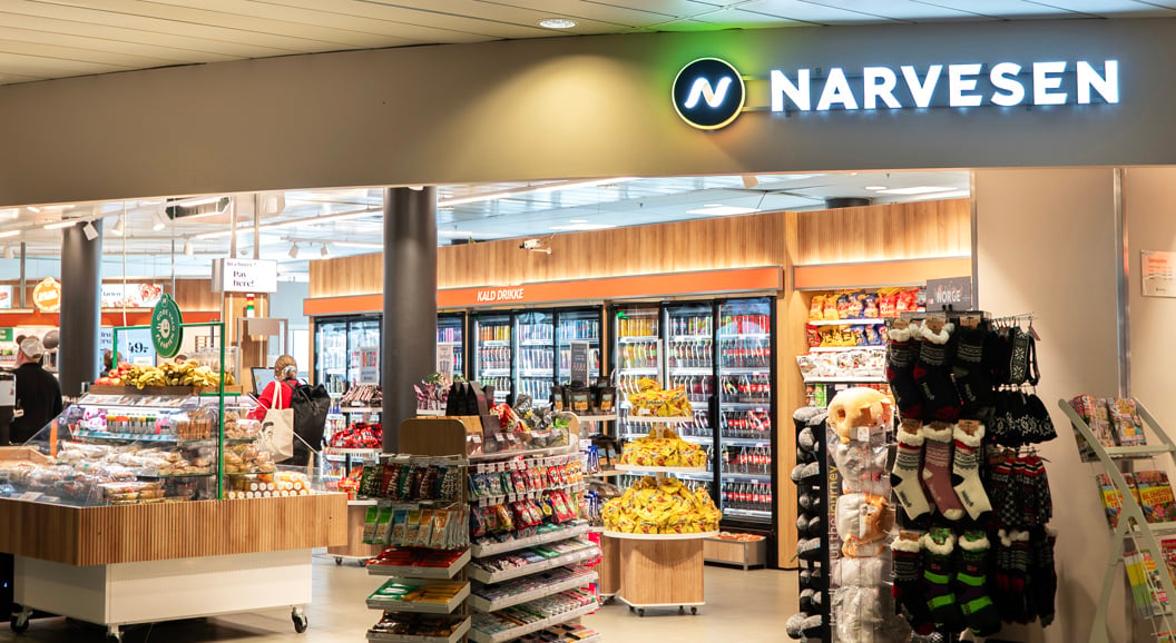 Narvesen butikkinteriør på en flyplass, med et utvalg av snacks, drikkevarer, magasiner og reiseartikler, med folk som ser seg rundt nær sitteområdene