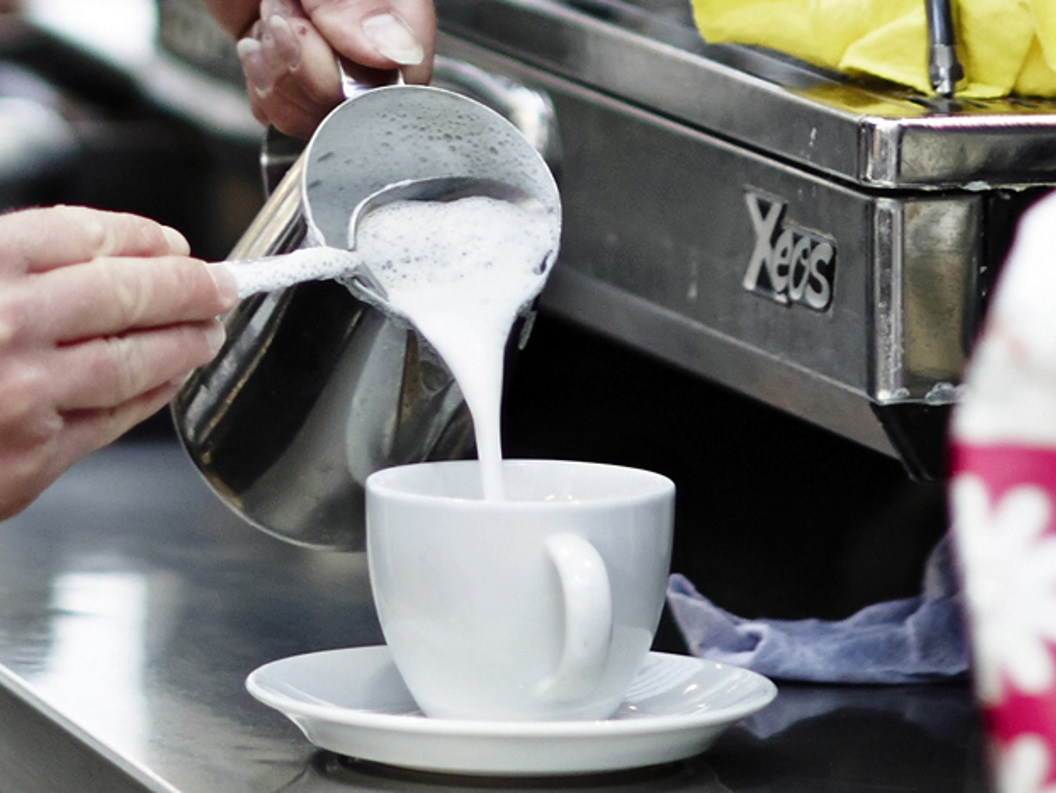Barista pouring steamed milk into a white coffee cup next to an espresso machine in a coffee shop