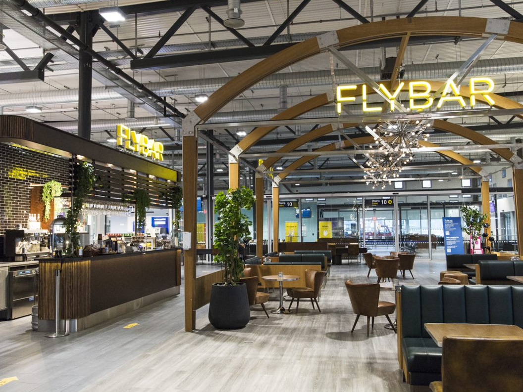 Flyplassbar med moderne interiør, lyst miljø med trepanel, grønne planter og sittegrupper i skinn. Puptre med opplyst skilt der det står "Flybar