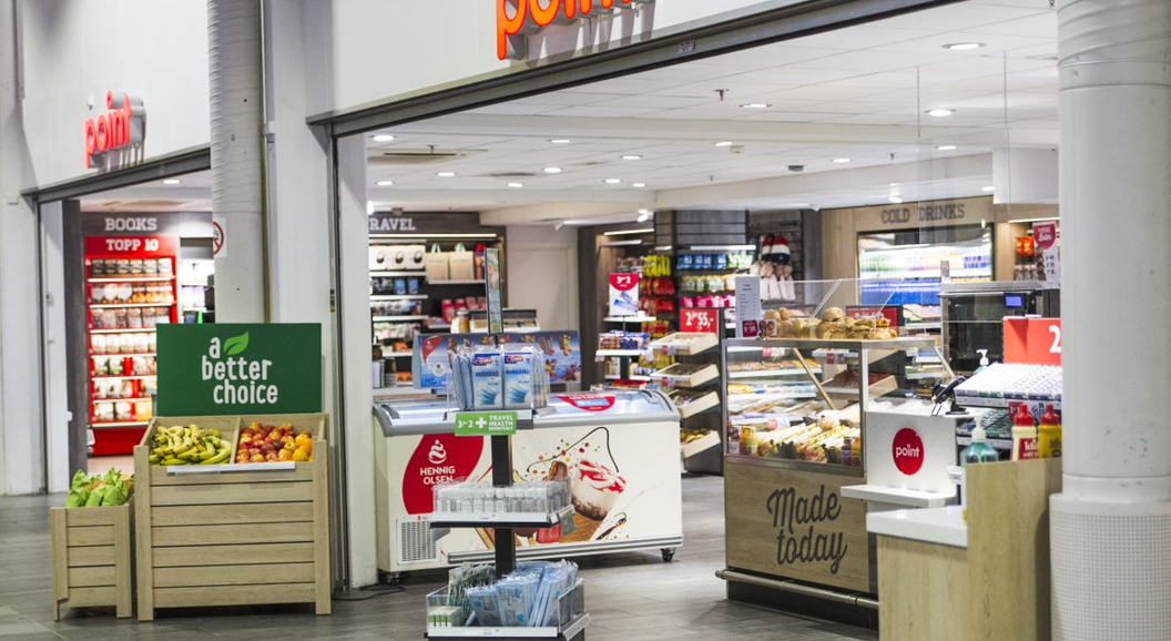 Inngang til en nærbutikk med utstilling av fersk frukt, magasiner, kalde drikker og en delikatesseavdeling med skiltet "Laget i dag". Ulike produkter er tilgjengelige for kunder