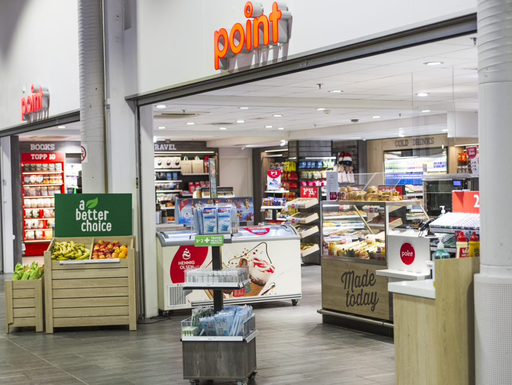 Butikkinteriør med mat og drikkevarer, frukt og snacks, hyller fulle av varer, og ein kaffimaskin. Skiltet "a better choice" framhevar sunne val