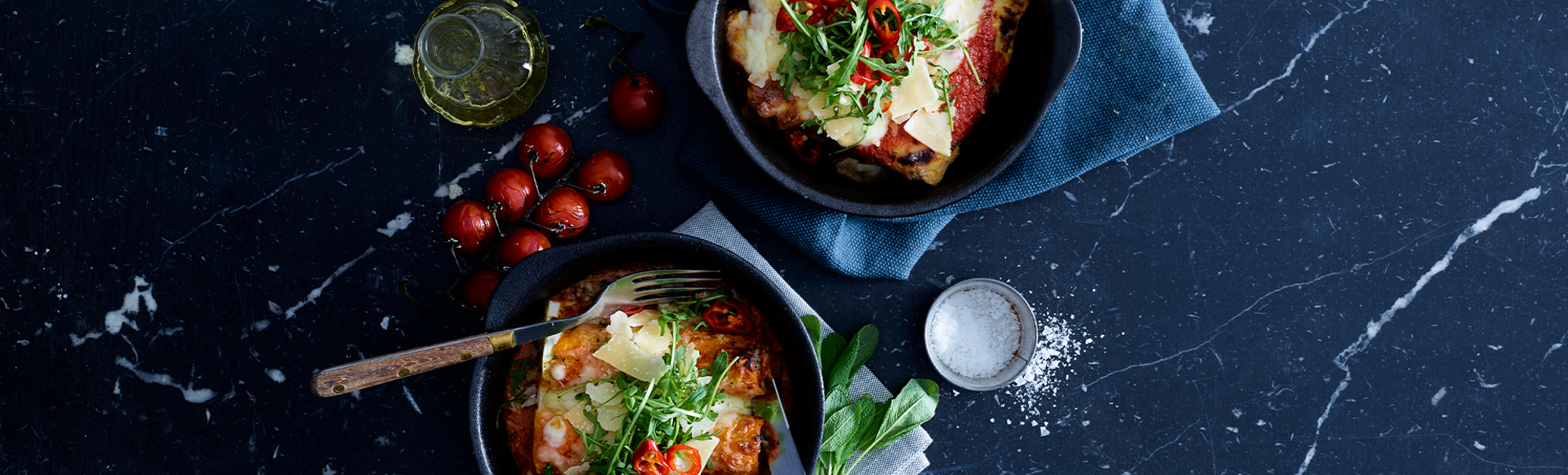 To skåler med lasagne toppa med urter og røde chili på mørk marmorbord, med småtomater, olivenolje og salt