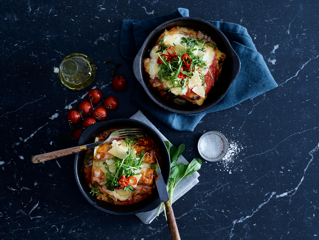 Two servings of lasagna in dark bowls on a marble surface, garnished with fresh greens and chili slices, accompanied by a small bottle of olive oil, cherry tomatoes, and a bowl of salt