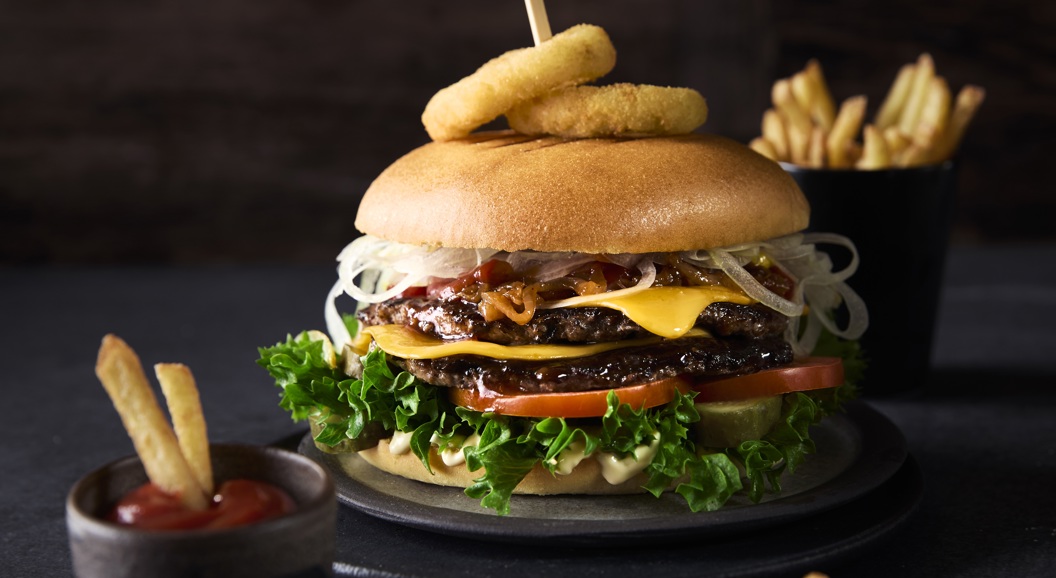 Deilig gourmetburger med salat, tomater, ost og løkringer på en bolle, pyntet med et spyd, akkompagnert av en side med pommes frites og ketchup