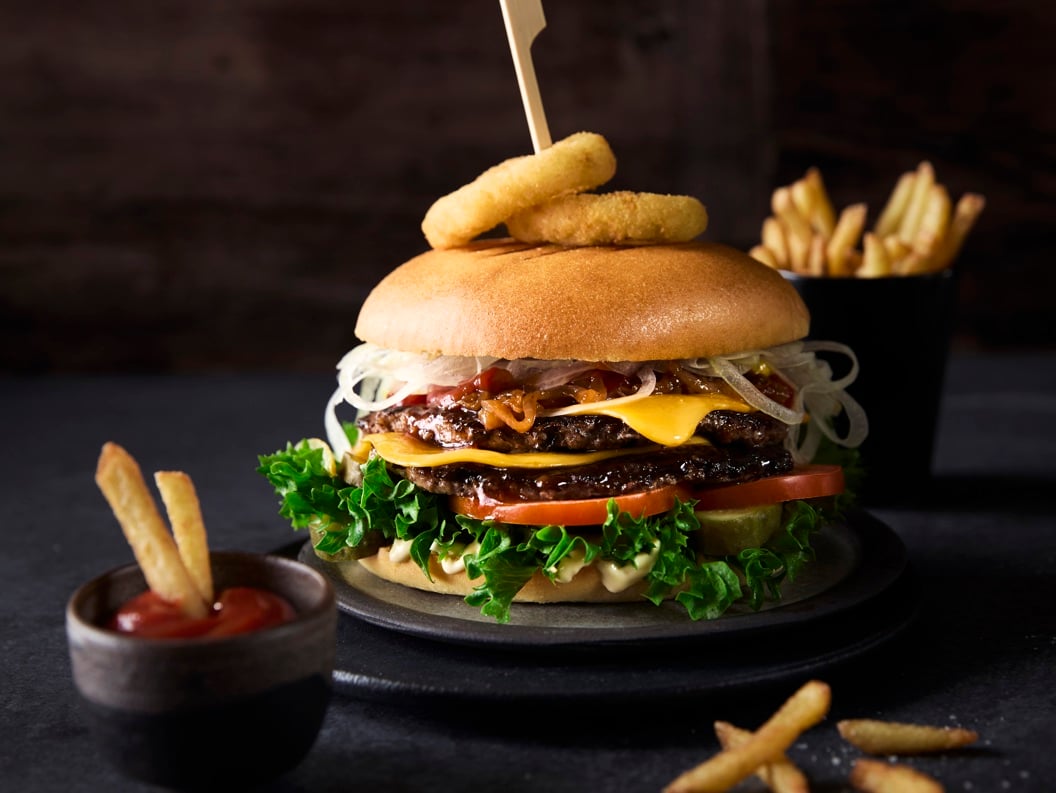 Deilig gourmetburger med salat, tomater, ost og løkringer på en bolle, pyntet med et spyd, akkompagnert av en side med pommes frites og ketchup