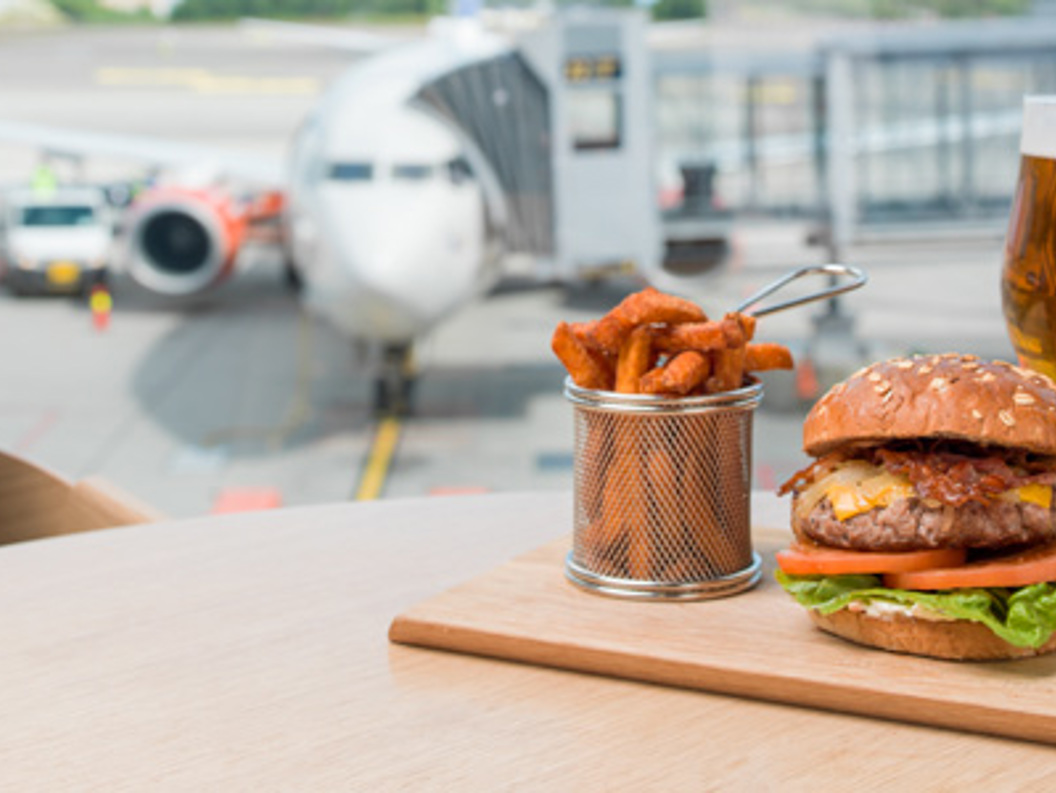 Gourmet burger with fries and a glass of beer on a table overlooking an airport runway with a parked airplane in the background