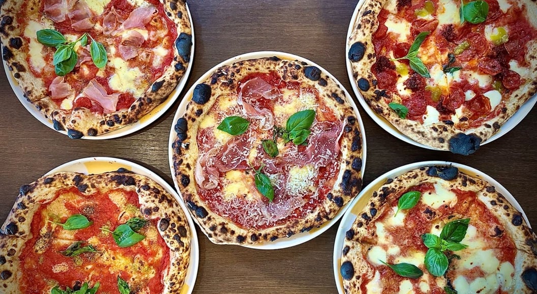 Fem håndverks-pizzaer med ulike toppinger inkludert basilikum, ost og spekemat på et trebord
