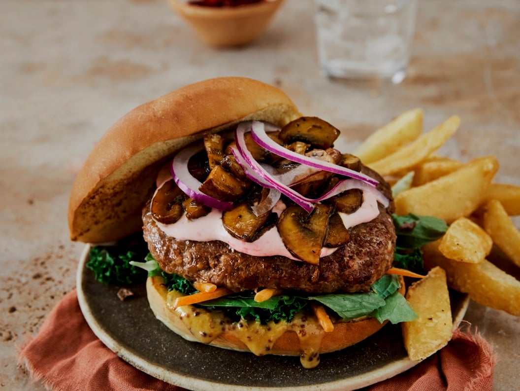 Saftig burger med rødløk, sopp, salat og dressing, servert med pommes frites på eit grått fat, ved sidan av glas med vatn
