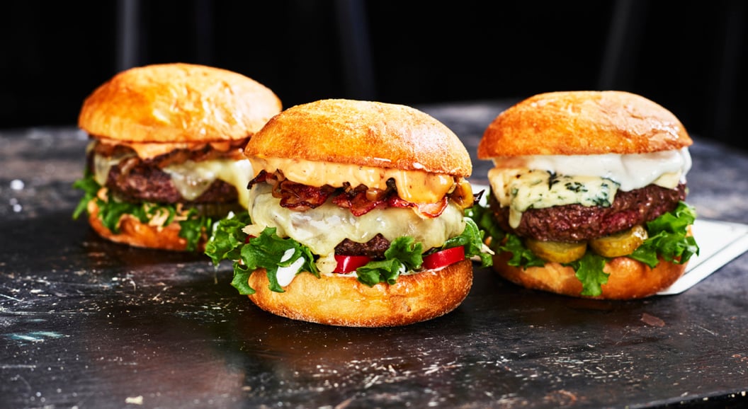 Three gourmet burgers with various toppings, including lettuce, cheese, pickles, and bacon, on a rustic black table