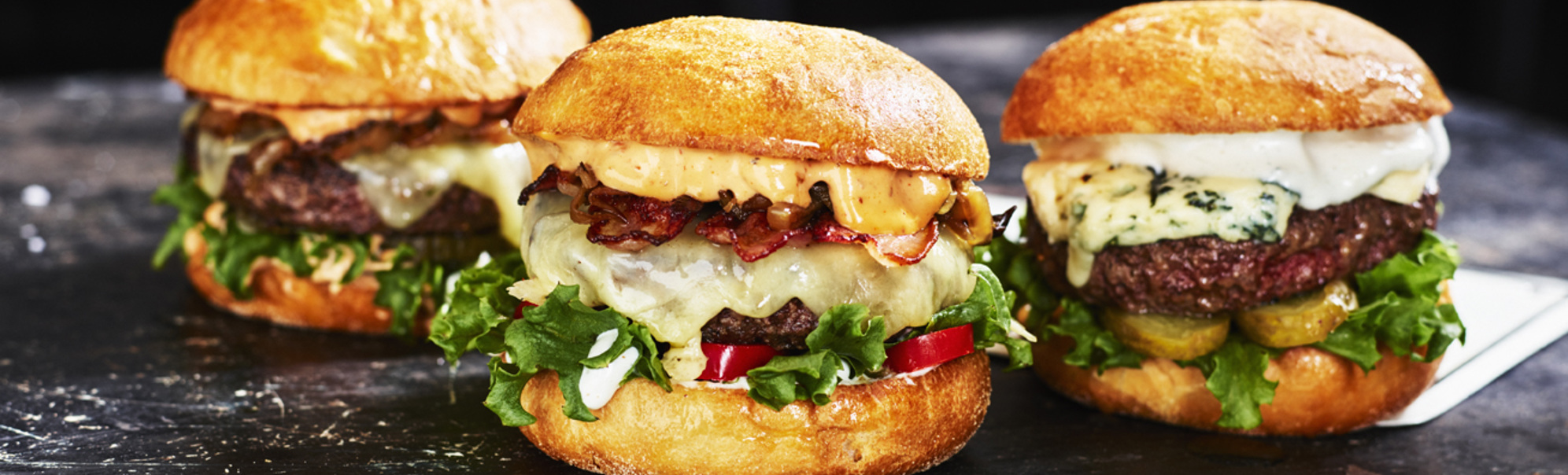 Tre gourmetburgere med ulike toppinger, inkludert salat, ost, pickles og bacon, på et rustikt svart bord