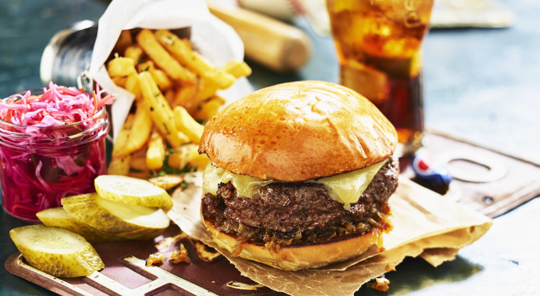 Juicy cheeseburger with melted cheese, accompanied by crispy French fries, sliced pickles, and coleslaw, with a glass of iced cola on a rustic table setting