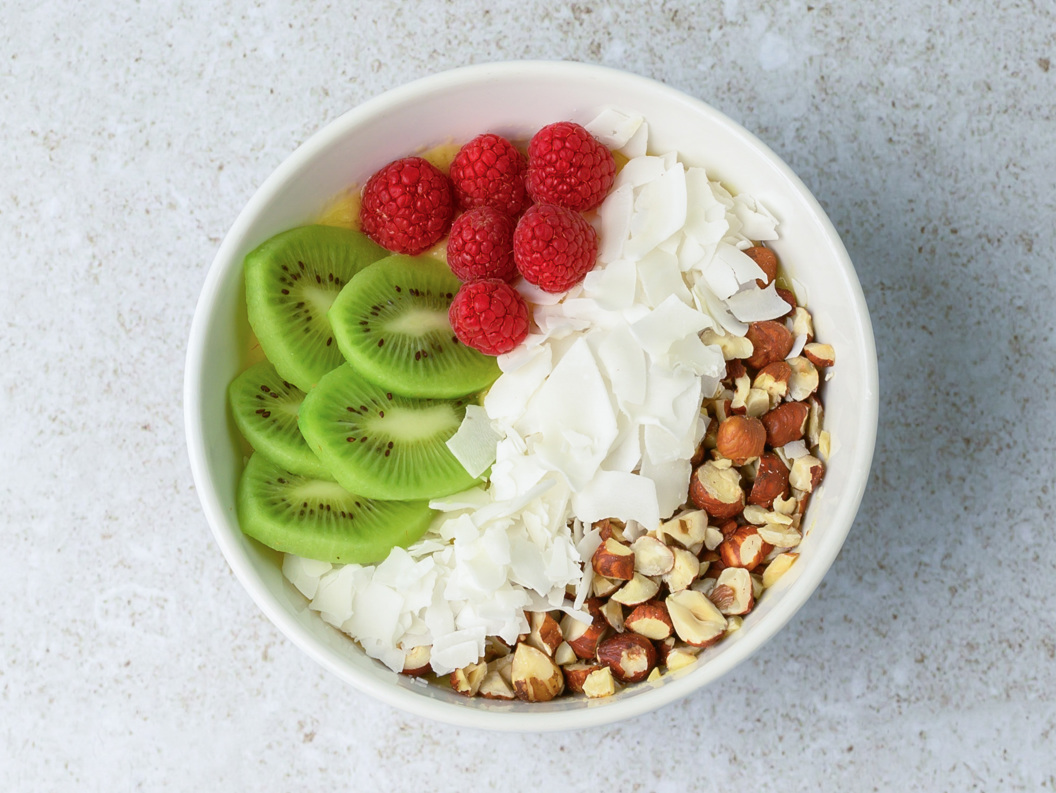 Yoghurtbolle med skiver av kiwi, bringebær, kokosflak og nøtter på ein lys overflate