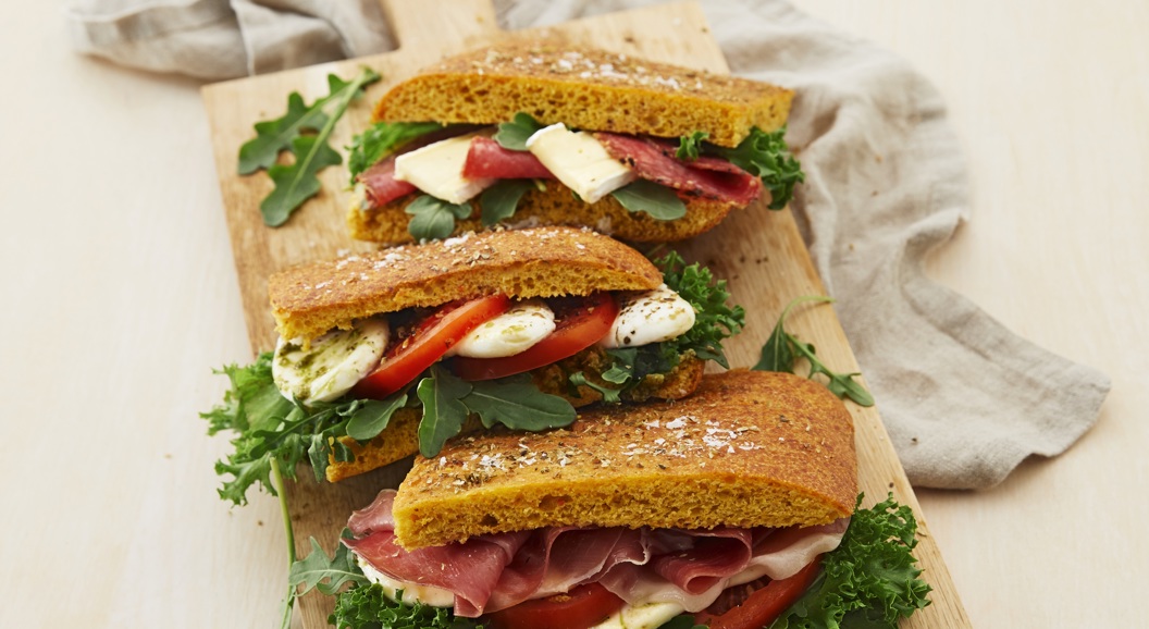 Three gourmet sandwiches with prosciutto, cheese, tomato, and arugula on a wooden board, styled with a linen cloth