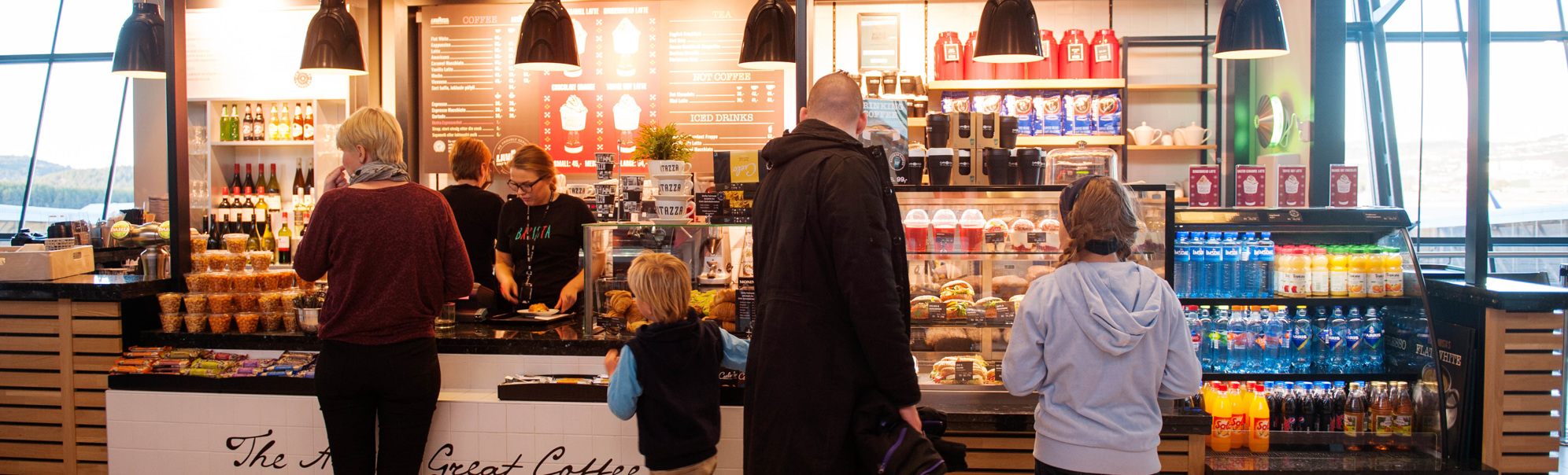 Skilt med teksten "Ritazza" på ein blå bakgrunn, omringa av grøne plantar i ein kafé