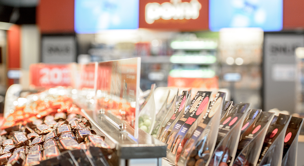 Godteriutstilling i en kiosk med sjokoladebarer og pakket godteri i lyse, organiserte rader. Uklart bakgrunn har drikkekjølere og reklameskilt