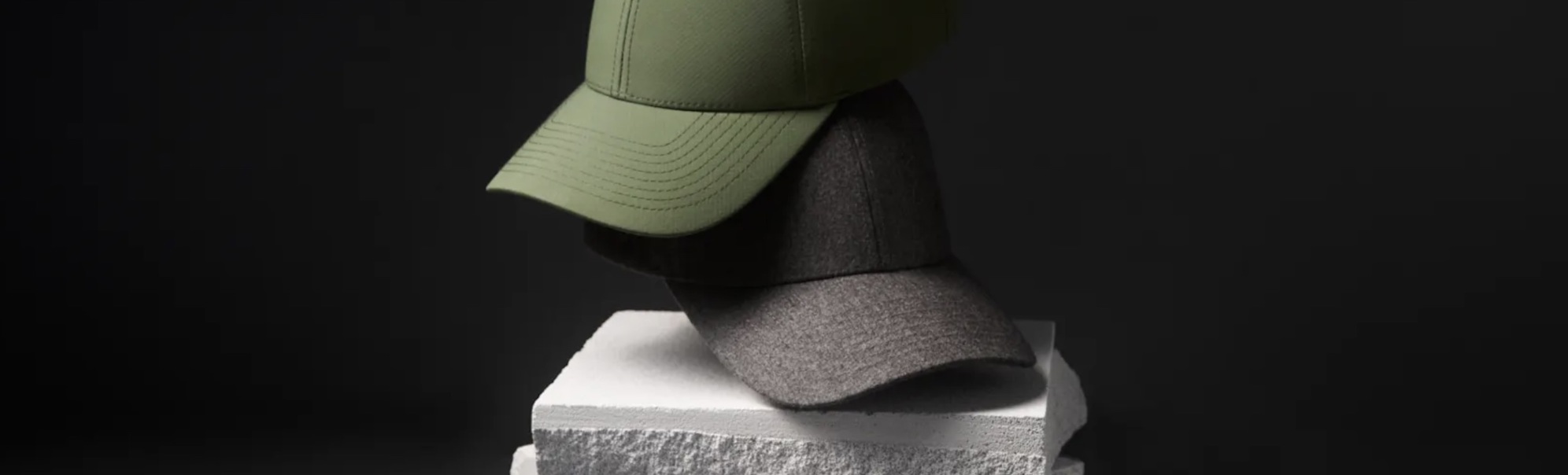 Two stylish caps, one green and one gray, are placed on stacked white concrete blocks with a dark background