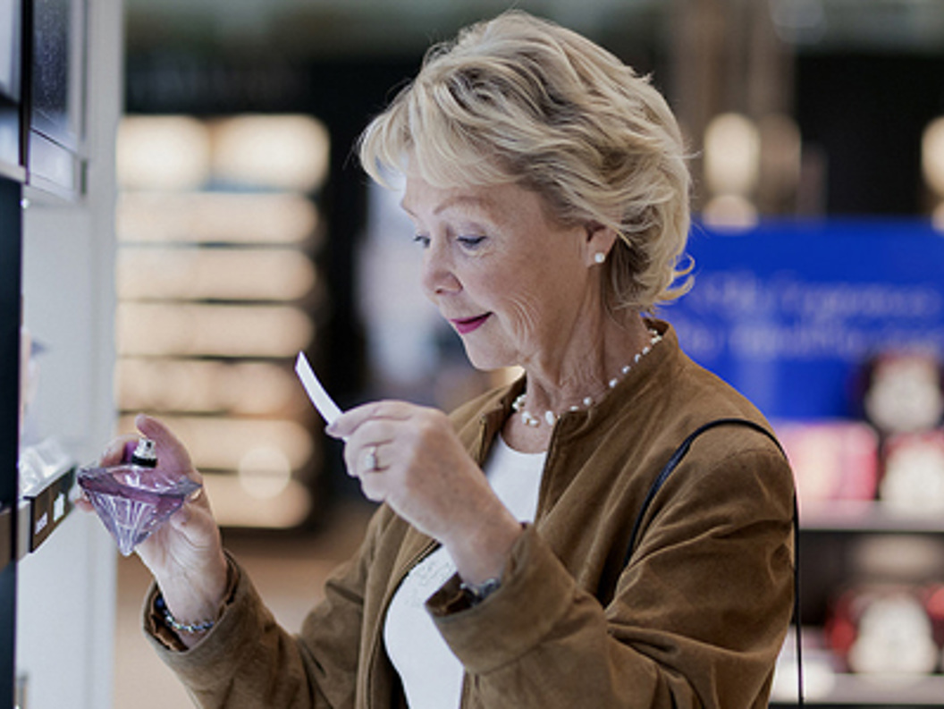 Senior woman testing perfume at a store, holding a fragrance strip while examining a perfume bottle, highlighting beauty shopping experience