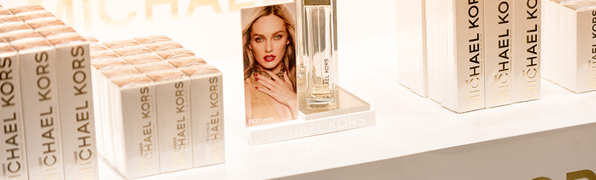 Visning av Michael Kors "Sexy Amber" parfyme med merkede emballasjebokser ryddig arrangert på en butikkhylle