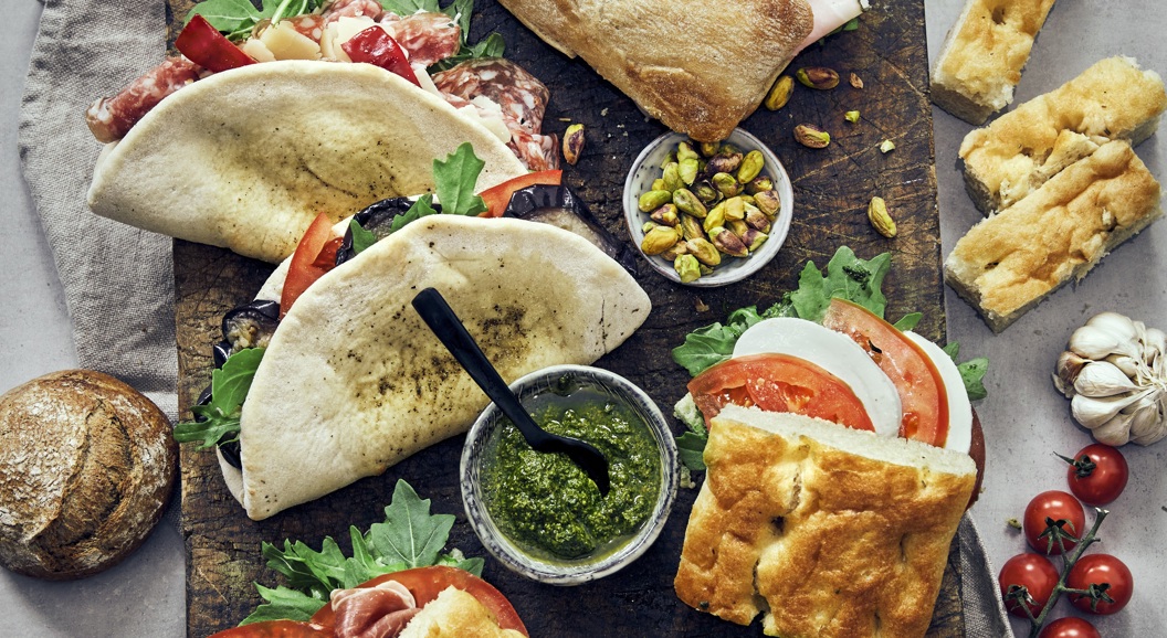 Italienske sandwicher med focaccia og ciabatta, fylt med spekemat, ost og grønnsaker. Serveres med pesto, aioli, nøtter og ferske cherrytomater. Matopplevelse på et trebord