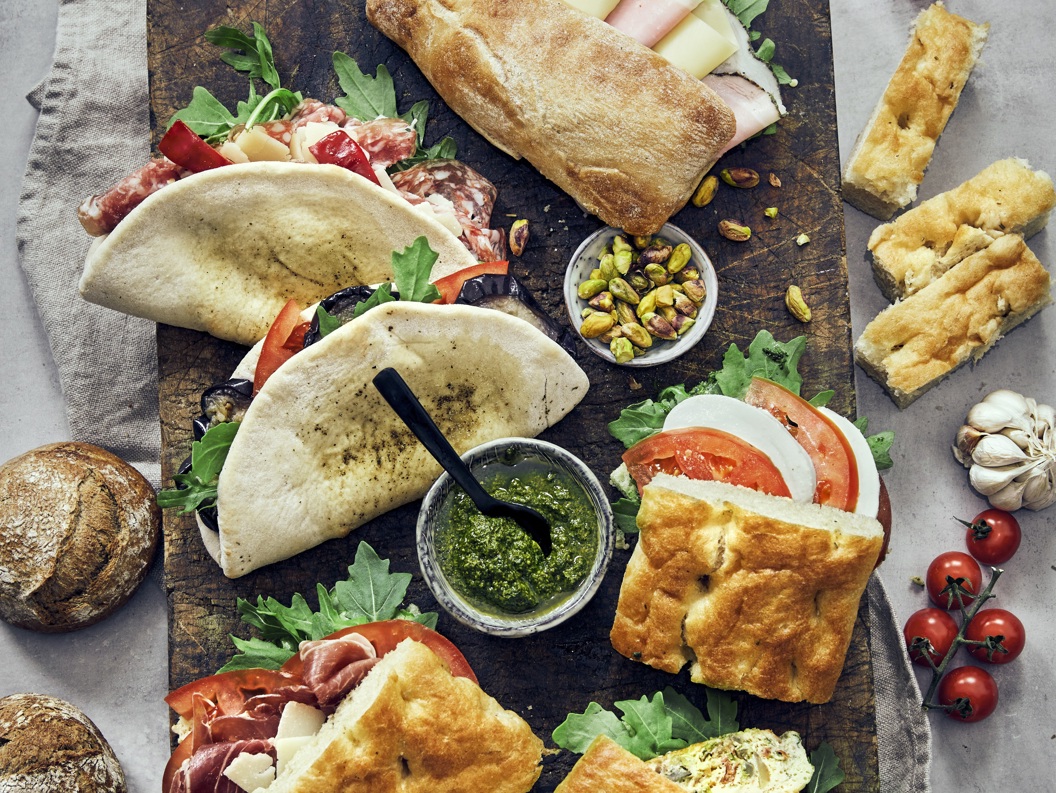 Italienske sandwicher med focaccia og ciabatta, fylt med spekemat, ost og grønnsaker. Serveres med pesto, aioli, nøtter og ferske cherrytomater. Matopplevelse på et trebord