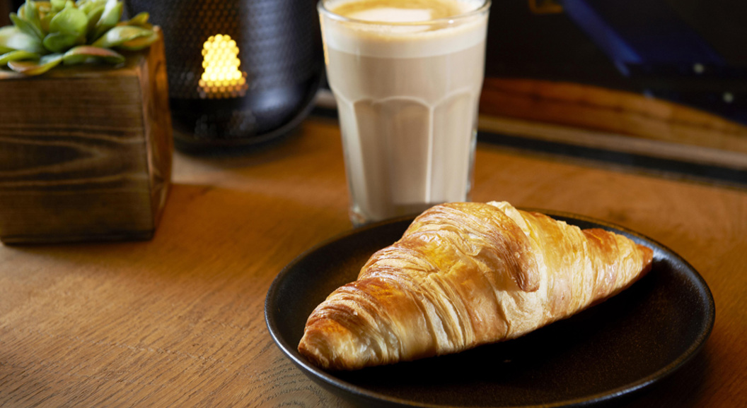 Fresh croissant on a black plate beside a glass of latte and a small potted plant on a wooden table
