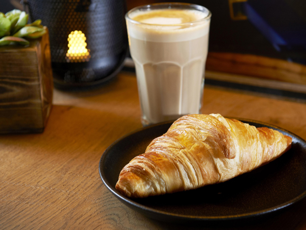 Fresh croissant on a black plate beside a glass of latte and a small potted plant on a wooden table