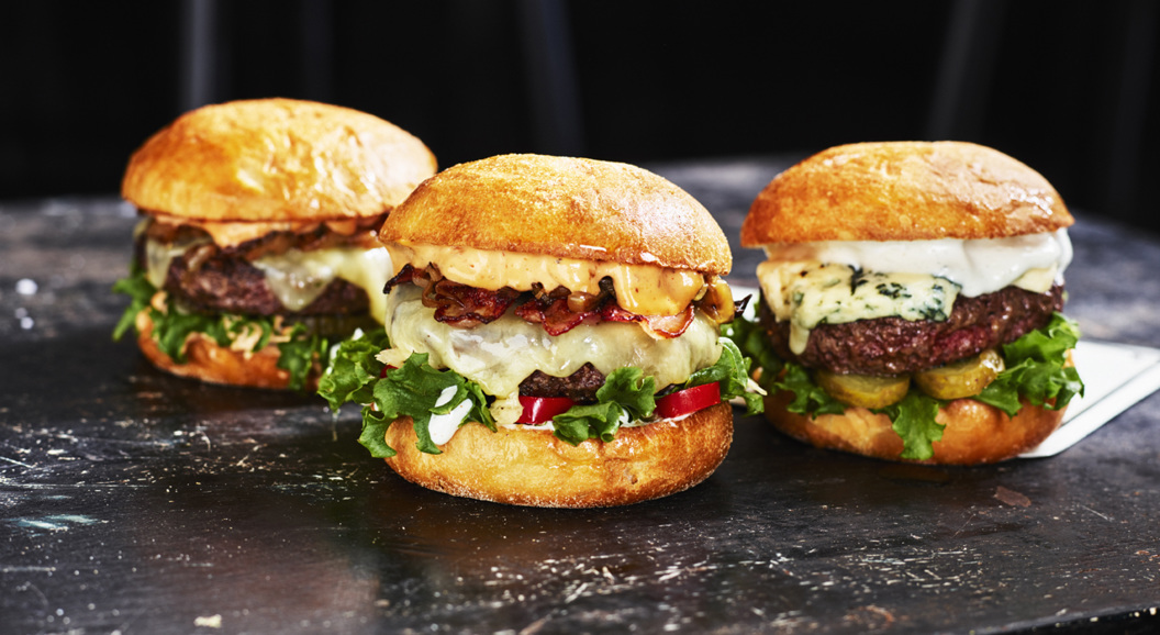 Tre saftige hamburgere med forskjellige pålegg på en mørk bordoverflate. Hver burger er fylt med smeltet ost, friske grønnsaker, og servert i luftige brioche-brød