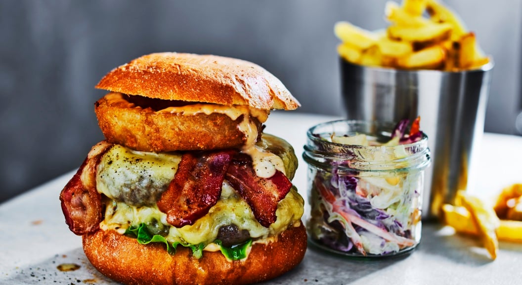 Gourmet cheeseburger with bacon, onion ring, and lettuce on a brioche bun, served with coleslaw and fries in a metal cup on a gray surface