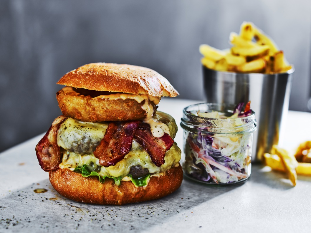 Gourmet cheeseburger with bacon, onion ring, and lettuce on a brioche bun, served with coleslaw and fries in a metal cup on a gray surface