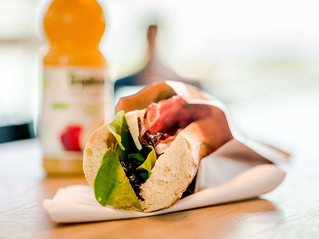 Freshly made sandwich with lettuce, bacon, and cheese on a wooden table, accompanied by a bottle of orange juice in the background