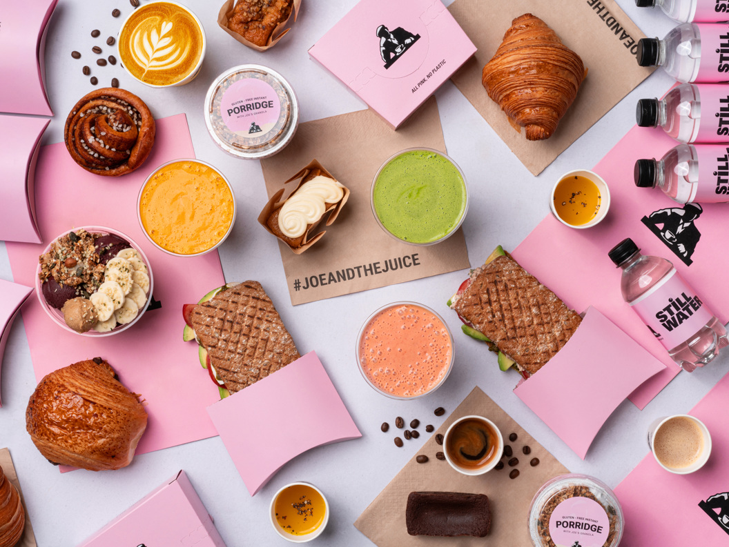 Assorterte Joe & The Juice-produkter, inkludert smørbrød, croissanter, grøt, juicer, açaí-boller og kaffe på en hvit bakgrunn med rosa emballasje og flasker med stille vann