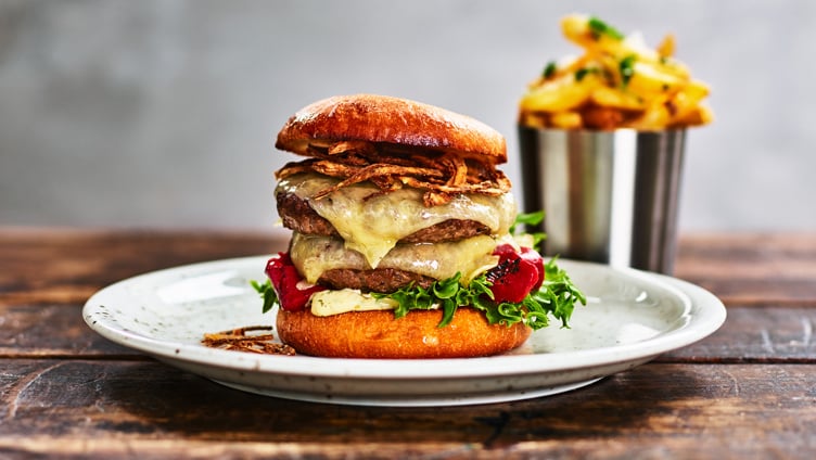 Gourmet cheeseburger with crispy onions, lettuce, and tomatoes on a toasted bun, served with a side of golden fries in a metal cup