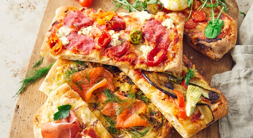 Fire rektangulære pizzastykker på trefjøl med ulike toppingar som spekeskinke, laks, burrata og grønsaker, servert med småtomatar og dippauser