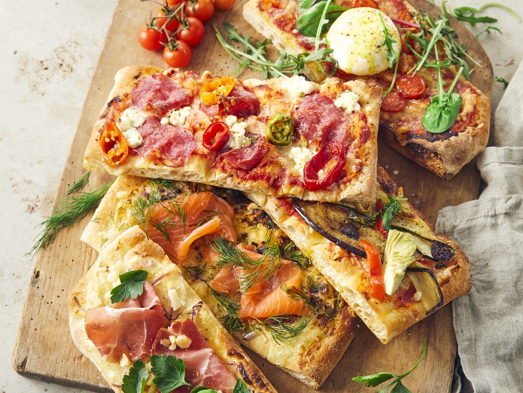 Fire rektangulære pizzastykker på trefjøl med ulike toppingar som spekeskinke, laks, burrata og grønsaker, servert med småtomatar og dippauser