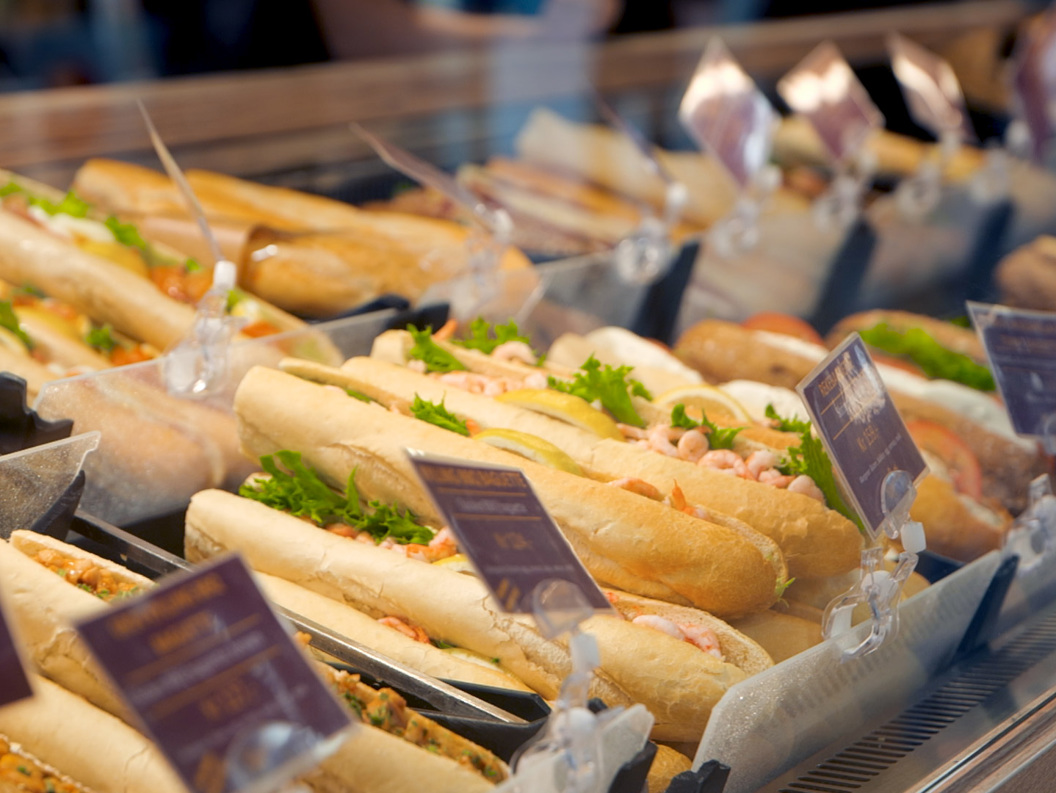 Nystekte baguettesmørbrød vist i en delikatedisk, med ulike fyllinger som reker og grønnsaker, med prislapper synlige i forgrunnen