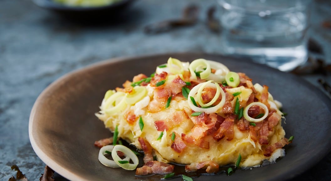 Potetmos toppa med sprø bacon og vårlauk på ein mørk tallerken, servert på eit rustikt bord