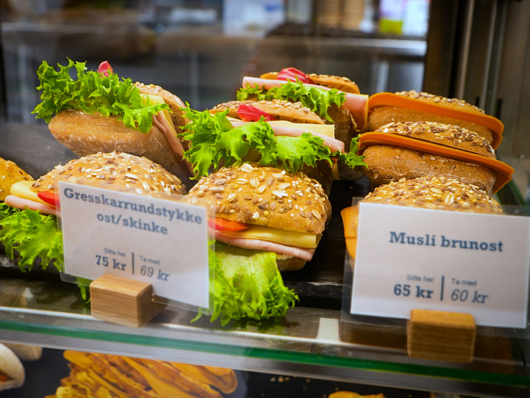 Assorterte smørbrød med salat, ost og kjøtt i en bakeridisk, med korn-toppede boller med synlige prislapper i norske kroner
