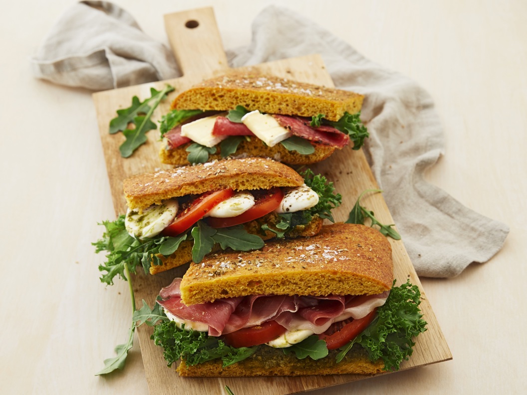 Three gourmet sandwiches with prosciutto, cheese, fresh greens, and tomatoes on a wooden board with a light cloth background