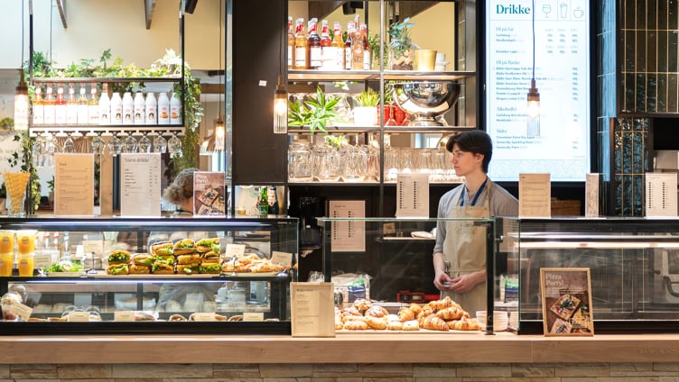 Moderne kaféinteriør med et utvalg av ferske bakverk, smørbrød og drikkevarer på utstilling; en ansatt står bak disken nær en pizzaovn