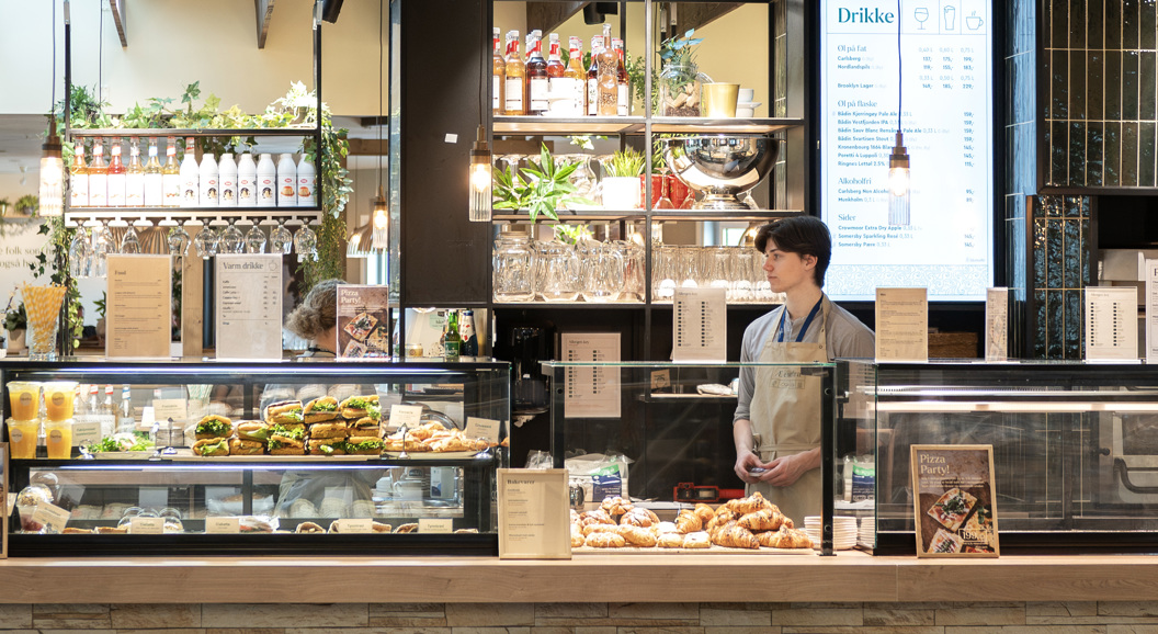 Modern café interior with a variety of fresh pastries, sandwiches, and beverages on display; a staff member stands behind the counter near a pizza oven