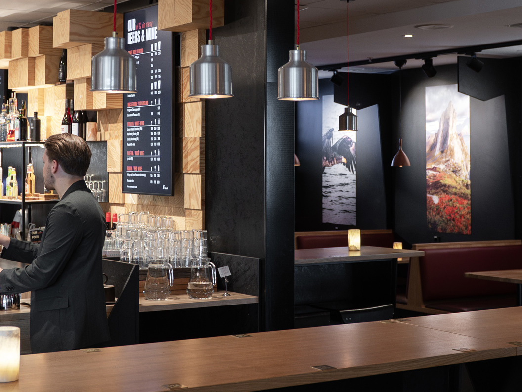 Koselig moderne kaféinteriør med en barista som tilbereder kaffe bak disken. Lokalet har moderne dekor, møbler i tre, hengende pendellamper, og innrammede landskapsbilder på veggene